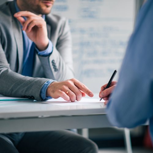 Low angle image of two men in formal wear sitting at the desk and and signing agreement.